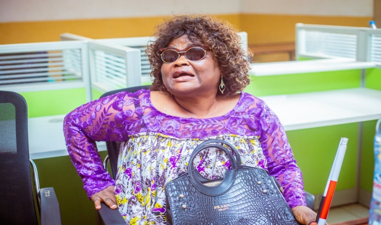 A woman in purple lace with dark glasses seated in the CAMAB meeting room, a white cane resting beside her.