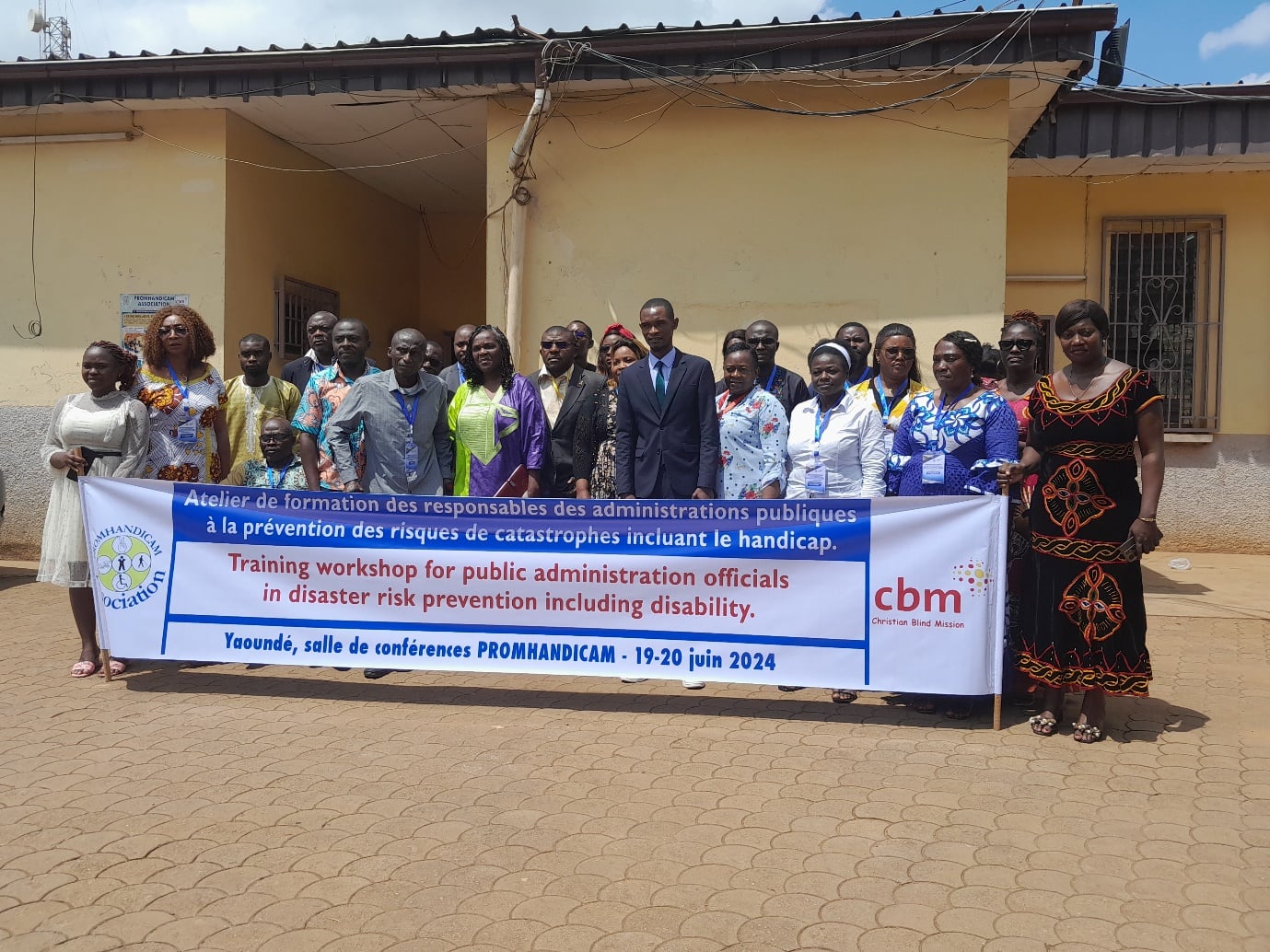 Group photograph outside a building behind a printed banner reading 'Atelier de formation des responsables des administrations publiques a la prevention des risques de catastrophes incluant le handicap / Training workshop for public...