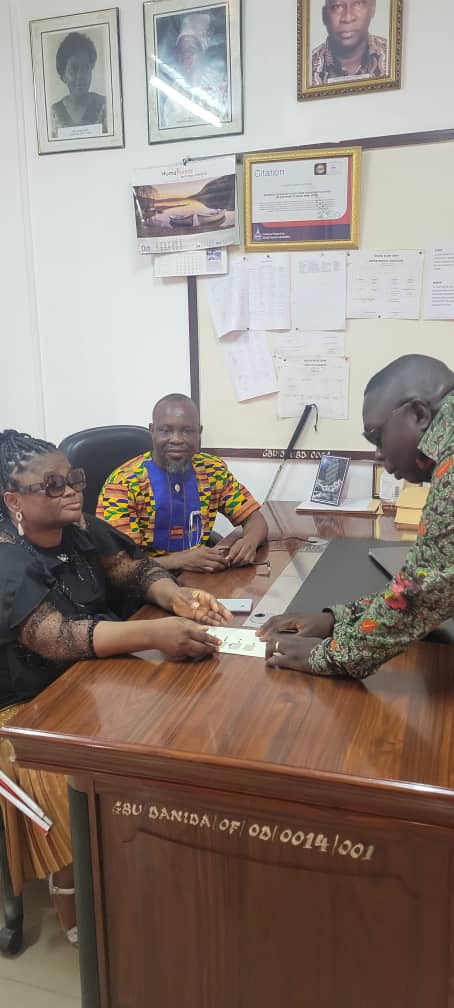 Office interior with framed certificates on the wall. Three people at a desk: a woman in dark glasses (blind) on the left, a man in a kente-pattern shirt centre, a third person right.