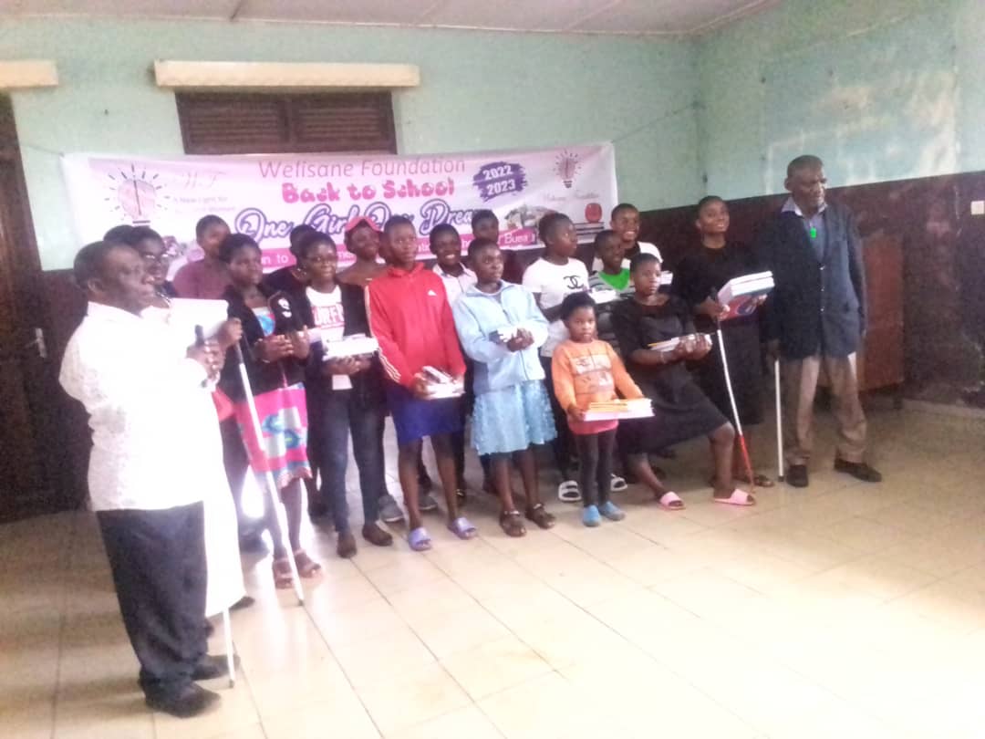 School-supplies handover under a printed banner reading 'Wefisane Foundation - Back to School - One Girl One Dream 2022/2023'. About fifteen children and adults hold exercise books and packets.