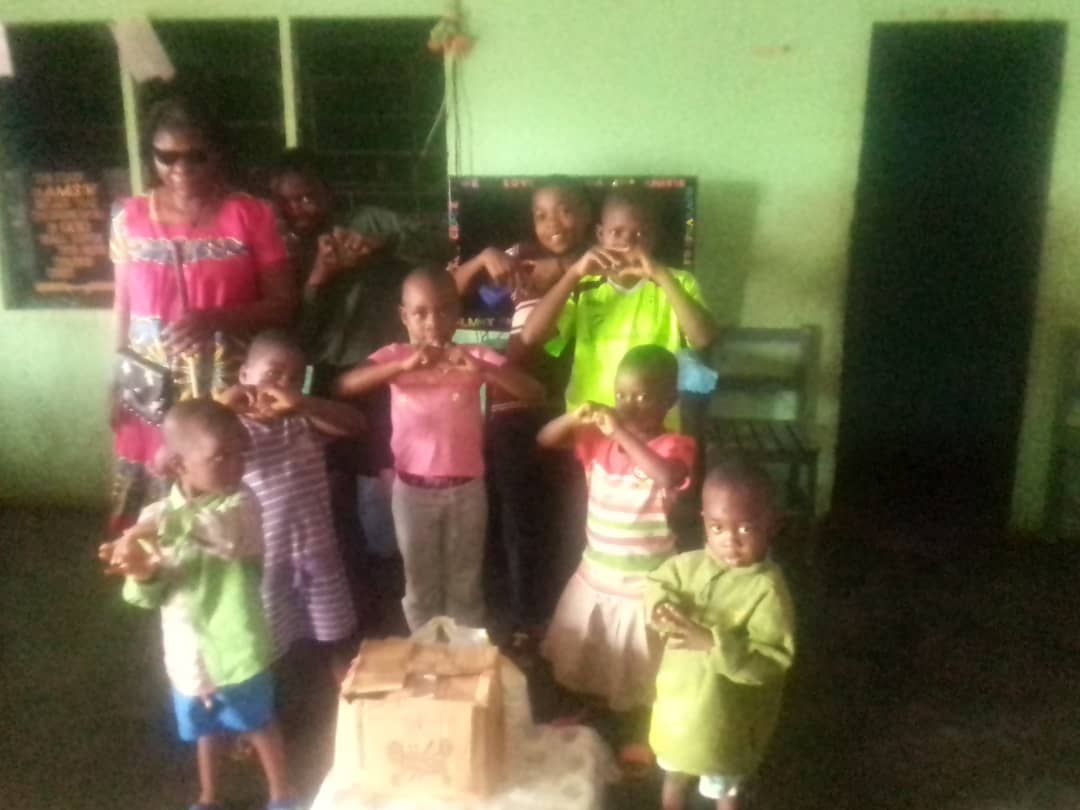 Classroom with dark green walls. Around eight children make heart shapes with their hands beside a woman in dark glasses and a pink top.