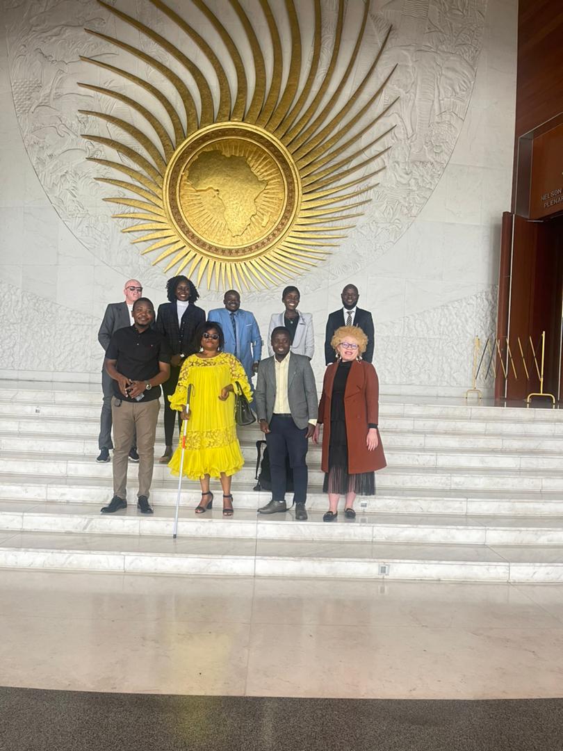 African Union headquarters, Nelson Mandela Plenary Hall. Nine delegates on the marble steps beneath the gold AU sunburst emblem; a woman in a bright yellow dress with a white cane stands front-centre.
