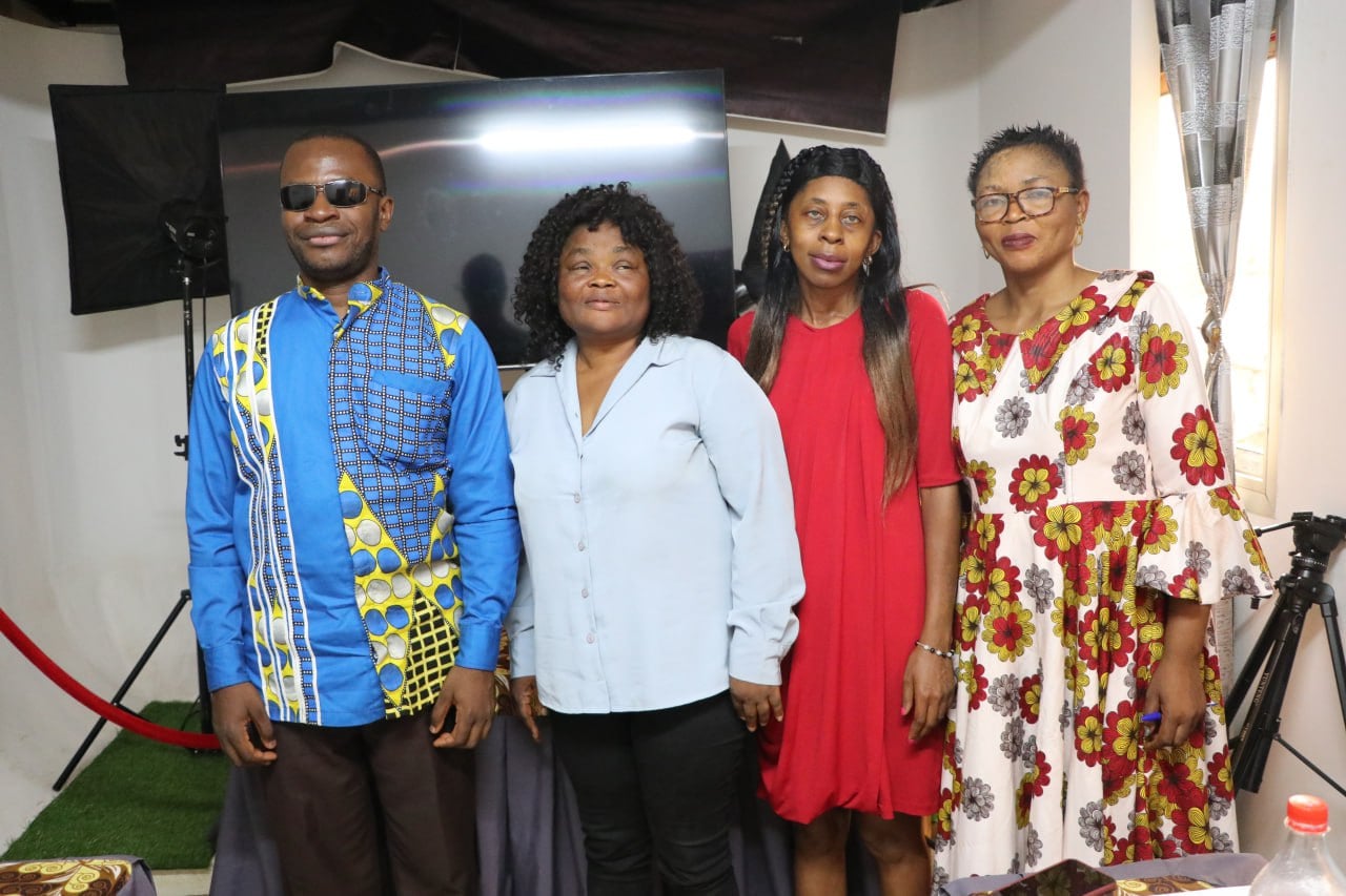 Television or photo studio: a man in blue Cameroonian print with dark glasses and three women stand in a row before a monitor. Softbox and tripod visible at the edges.