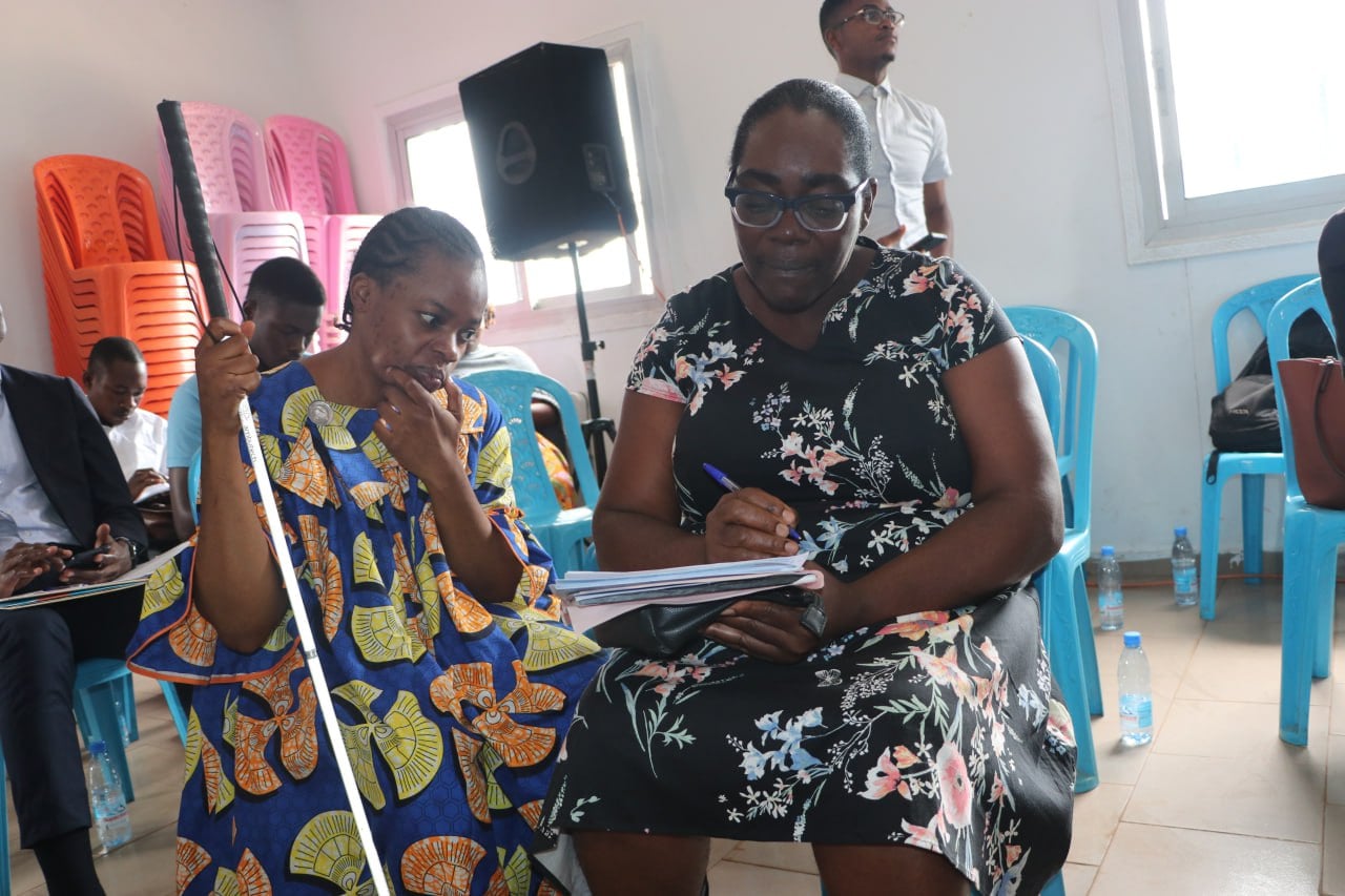 Workshop room with blue plastic chairs. A blind woman holding a white cane sits beside a woman in a floral dress who is writing notes on her behalf.