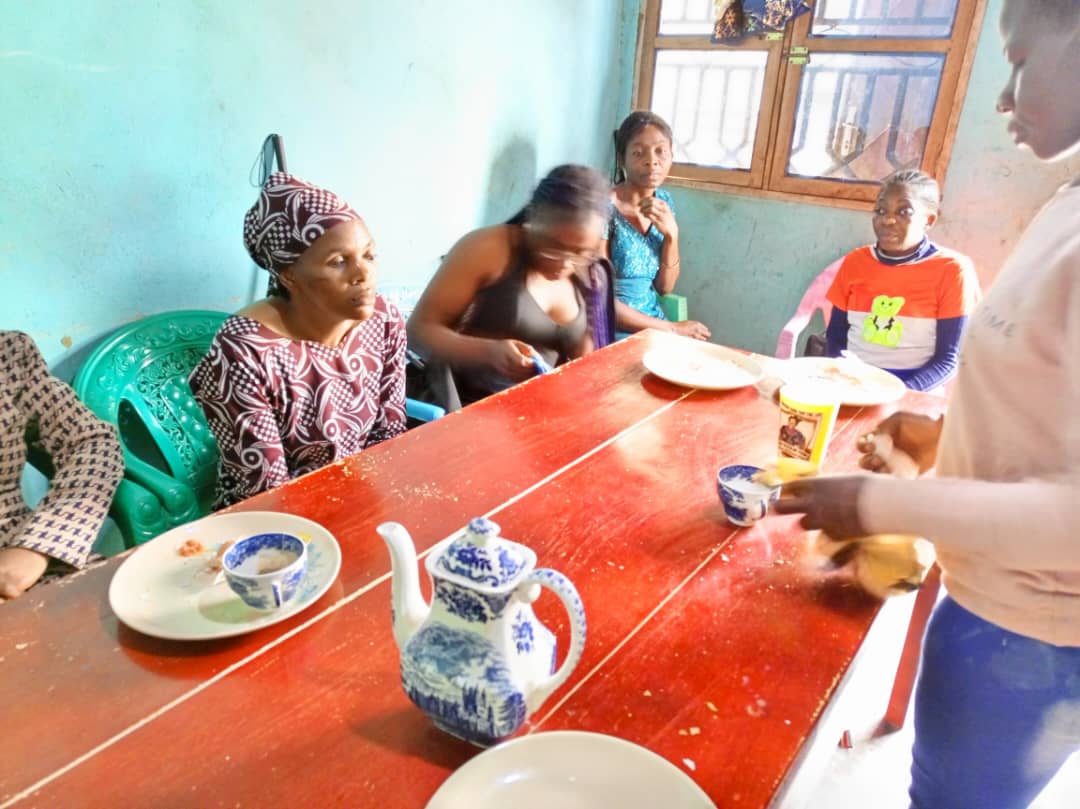 Community room with a red-painted table. Women seated on plastic chairs; tea being poured, plates and a blue-and-white teapot on the table.