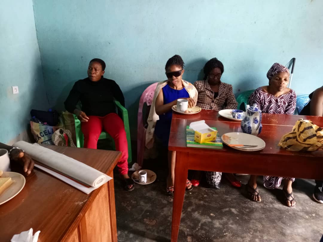 Room with the red table; women seated along the wall, one standing in red trousers. Teapot, plates and a tissue box on the table.
