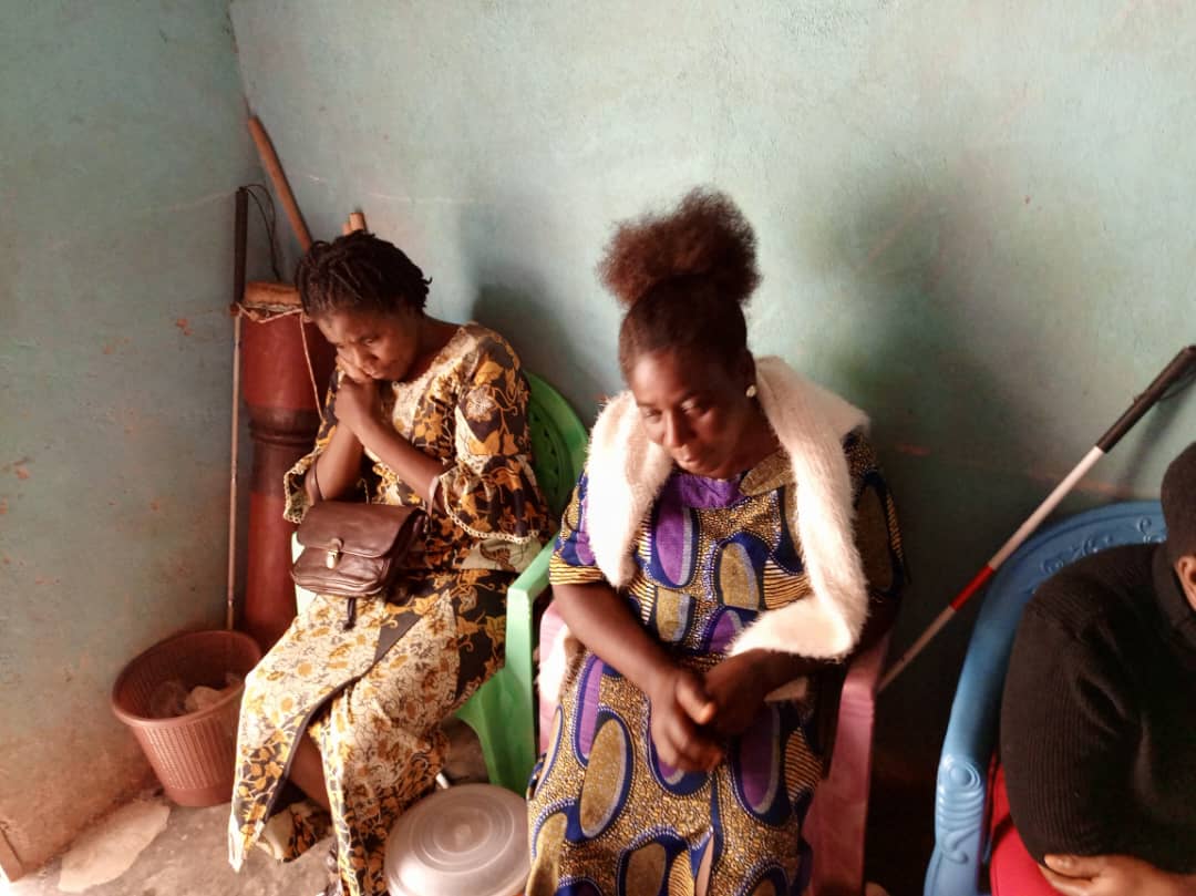 Two women seated in a corner, one in gold print, one with a fur-trimmed collar. A cane is visible at the right.