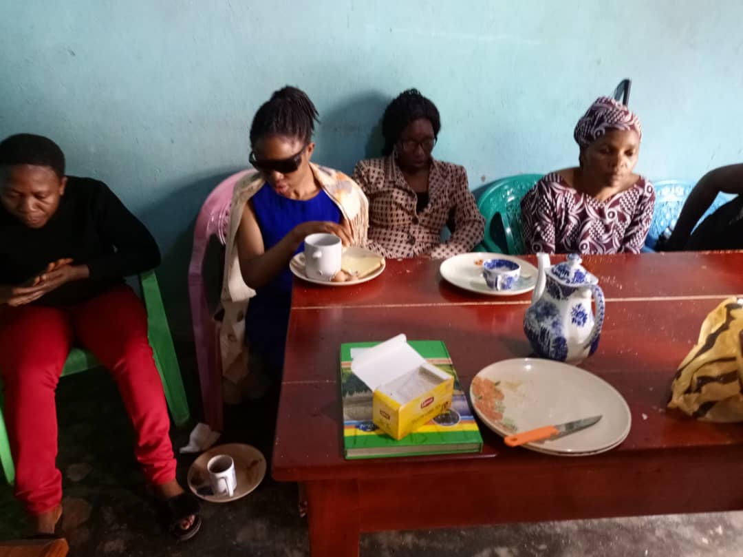 Women seated around the red table; one in dark glasses holds a cup and saucer. Tissues, a knife and plates on the table.