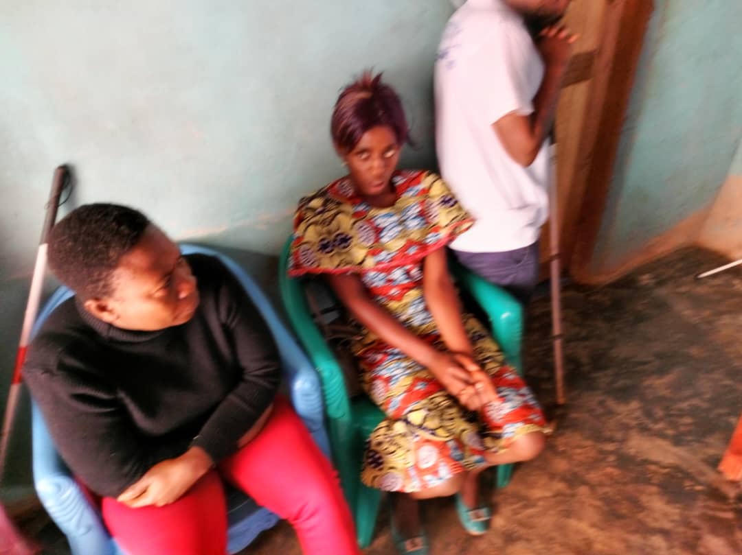 A woman in a bright print dress seated while another person bends in front of her; a cane and a third person by the doorway.