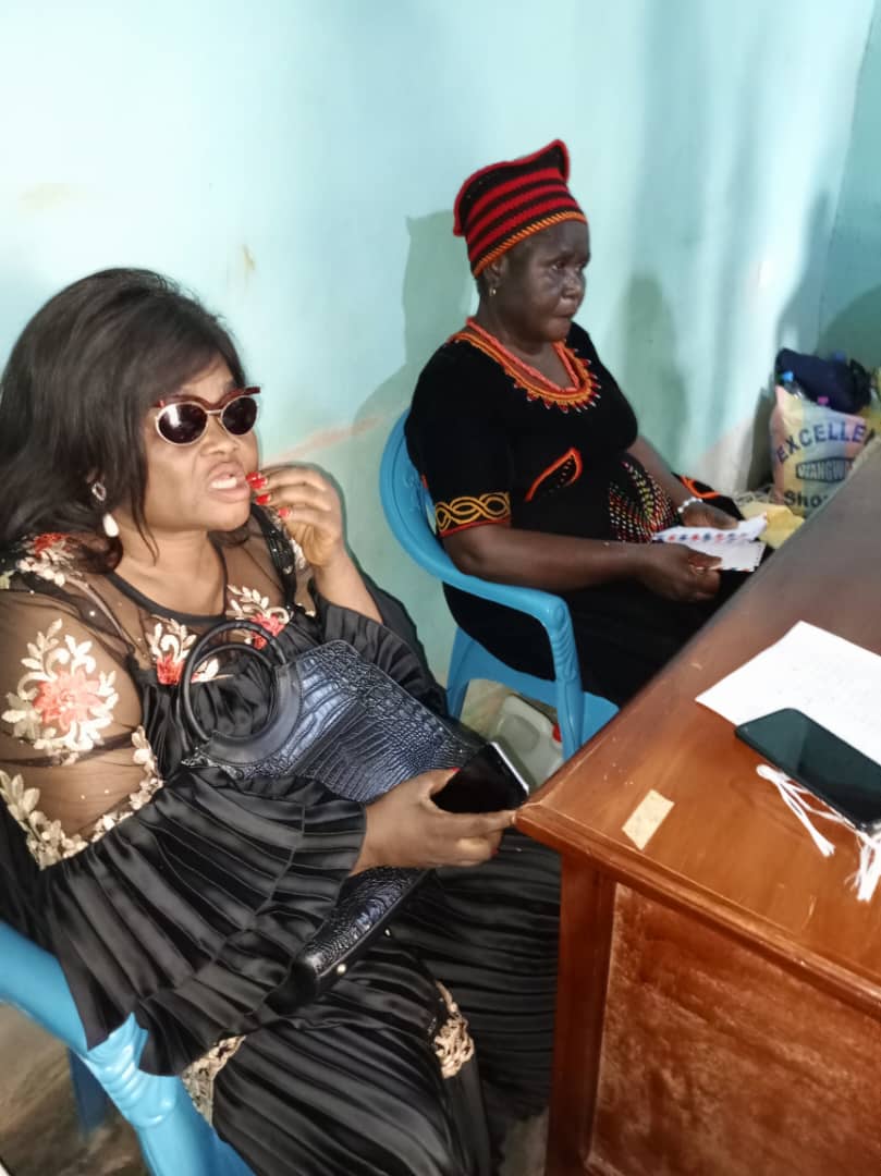 A woman in black lace with dark glasses at a desk beside a woman in a red Bamileke-style cap; papers between them.