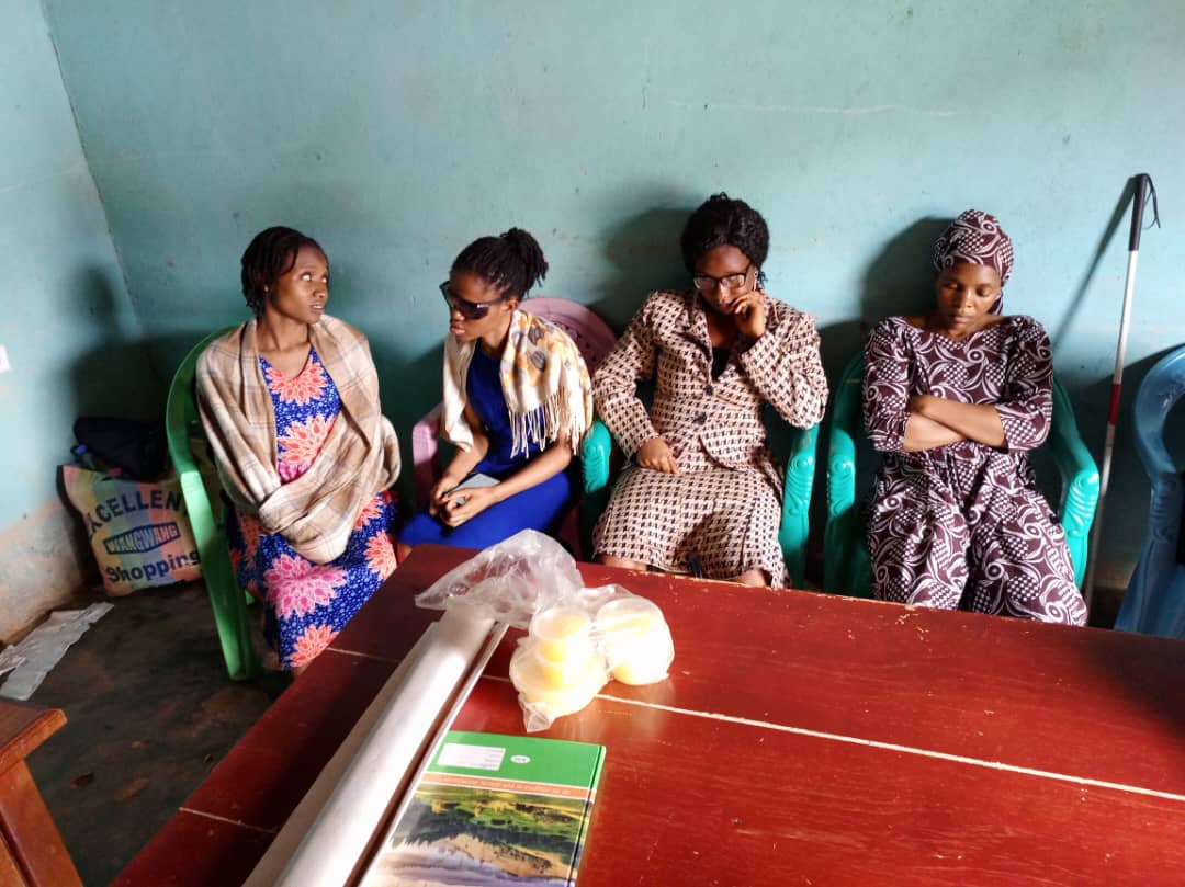 Four women seated in a row against the blue-green wall; a green booklet and a bag of bread on the red table.