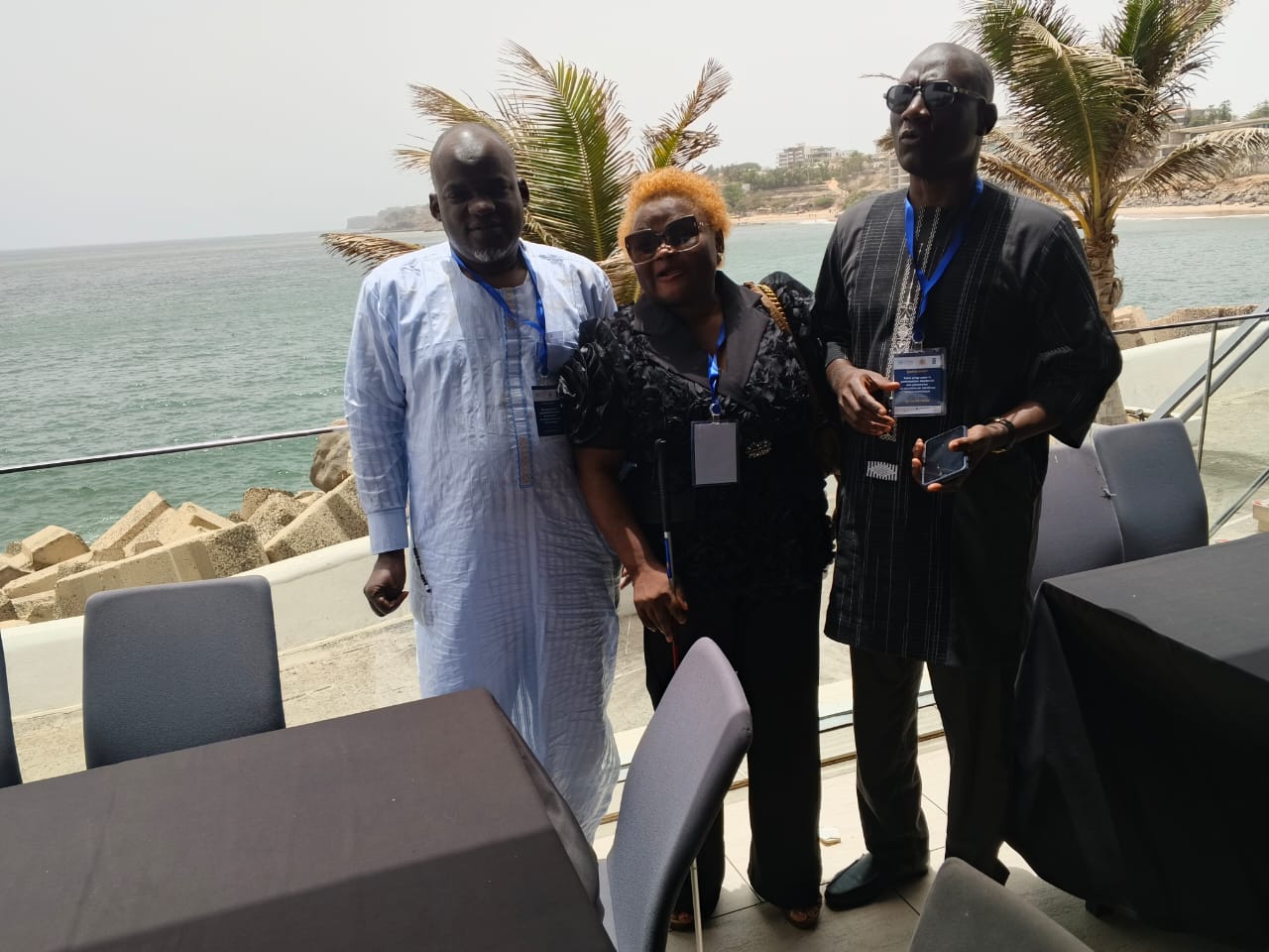 Seaside conference terrace (Dakar). Three delegates with conference lanyards stand together; the woman centre wears dark glasses and holds a white cane.