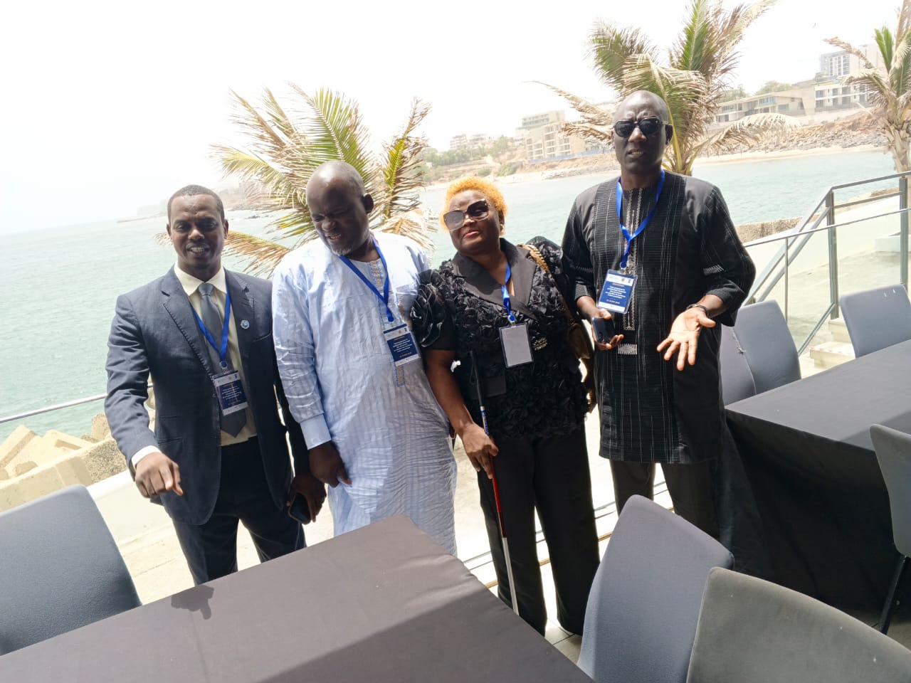 Seaside conference terrace (Dakar). Four delegates with lanyards in a row; the woman third from left wears dark glasses and holds a white cane.