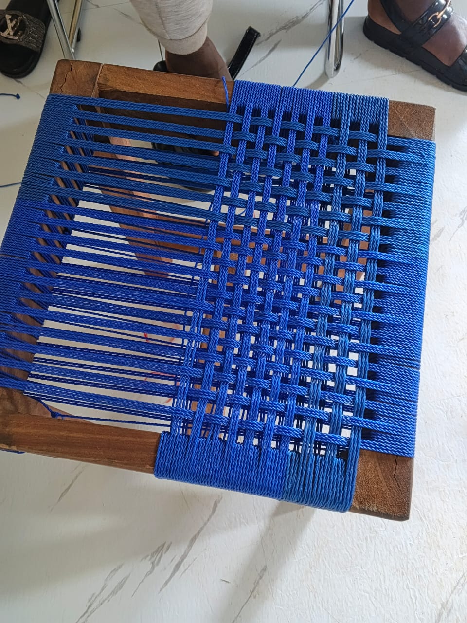 Close overhead detail of a wooden stool seat being woven with blue cord across the frame.