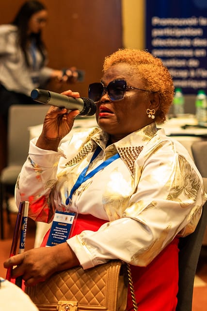A woman in a cream and gold outfit speaks into a hand-held microphone at a conference table.