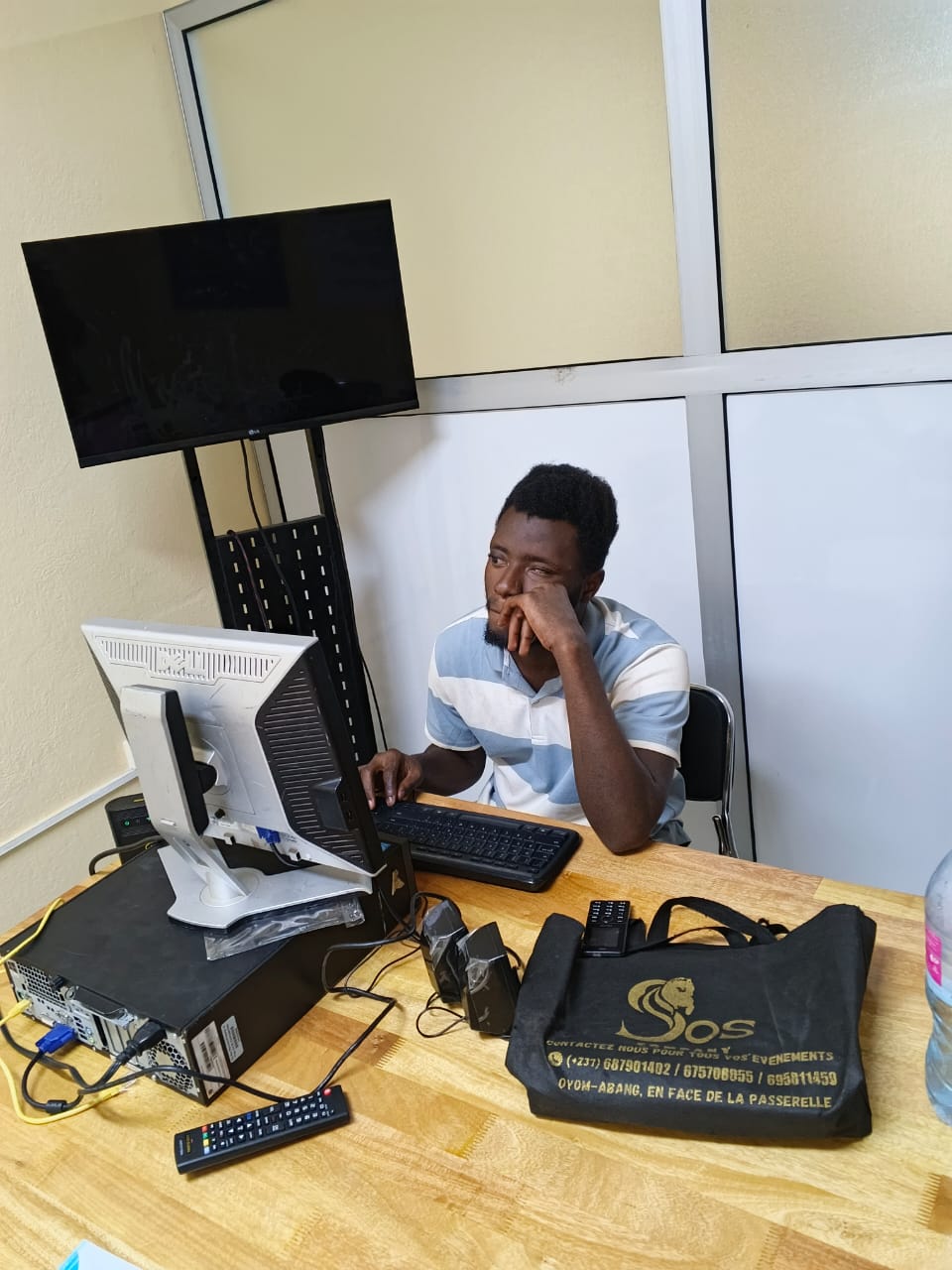 A young man works at a desktop computer in an office; a branded shoulder bag on the desk, monitor and keyboard in front of him.
