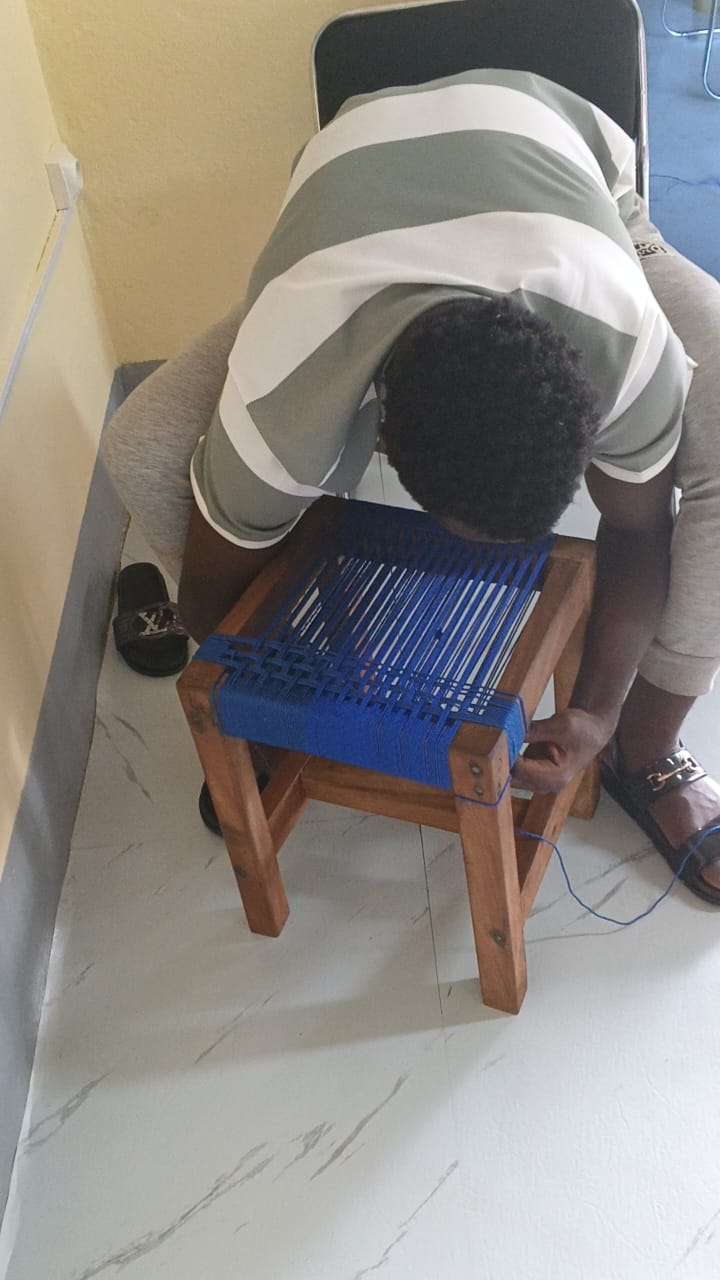 Overhead view of a man bent over a wooden stool, threading blue cord across the seat frame.