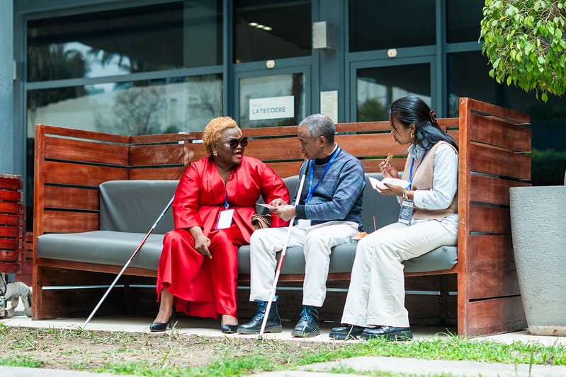 Outdoor bench at a conference venue: a woman in red with a white cane, a man with a white cane, and a woman in white taking notes, seated together in conversation.