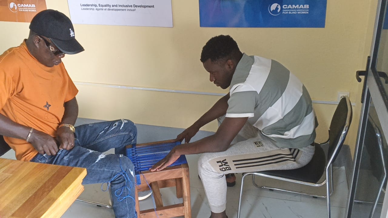 Two men seated in the CAMAB centre weaving a stool seat with blue cord; one wears dark glasses and a cap. CAMAB banners on the wall behind read 'Leadership, Equality and Inclusive Development' and 'CAMAB - Cameroonian Association for...