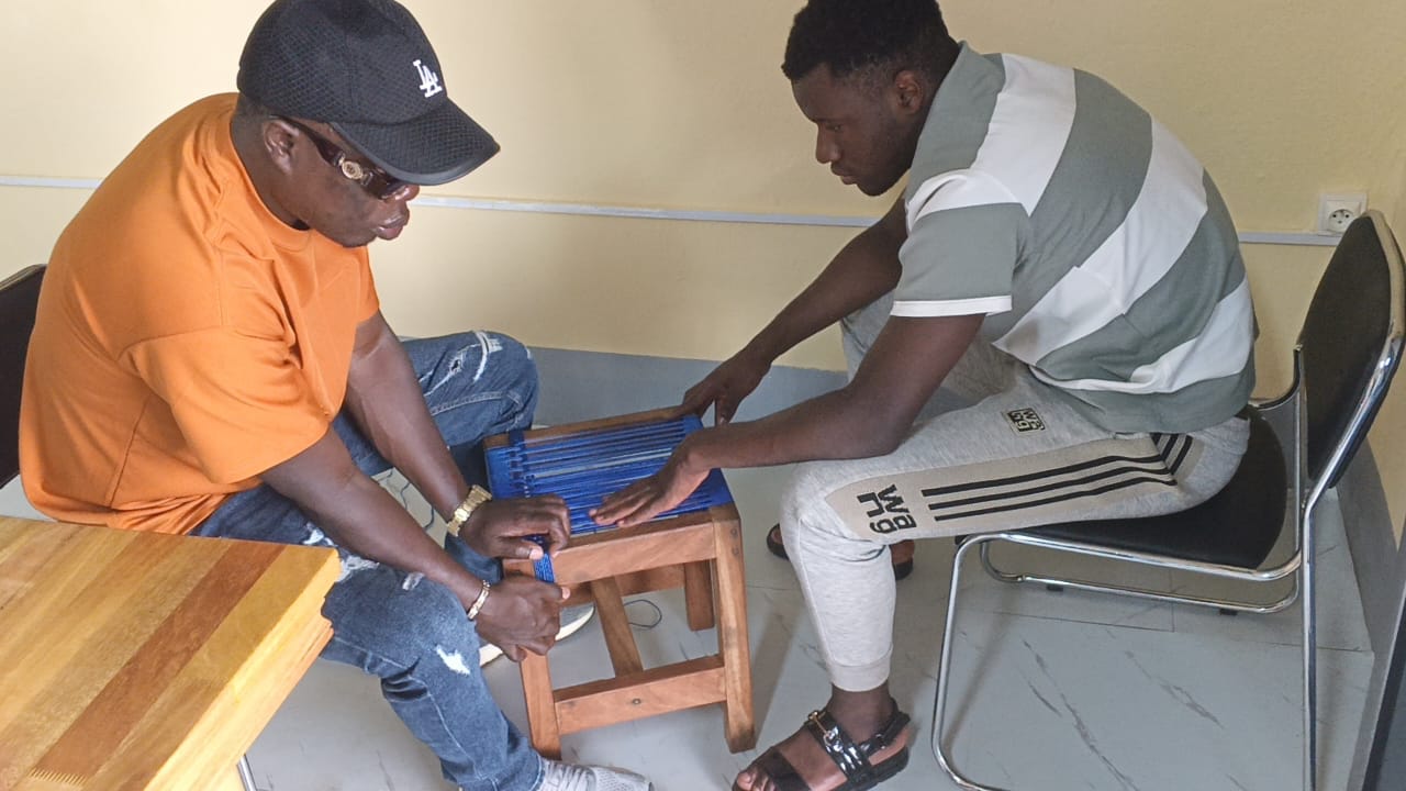 Two men seated in the CAMAB centre weaving a stool seat with blue cord; one wears dark glasses and a cap. CAMAB banners on the wall behind read 'Leadership, Equality and Inclusive Development' and 'CAMAB - Cameroonian Association for...