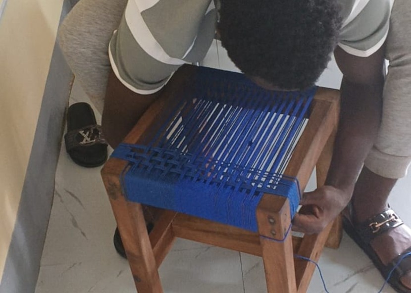 A learner weaving a stool seat with blue cord during handicraft training.