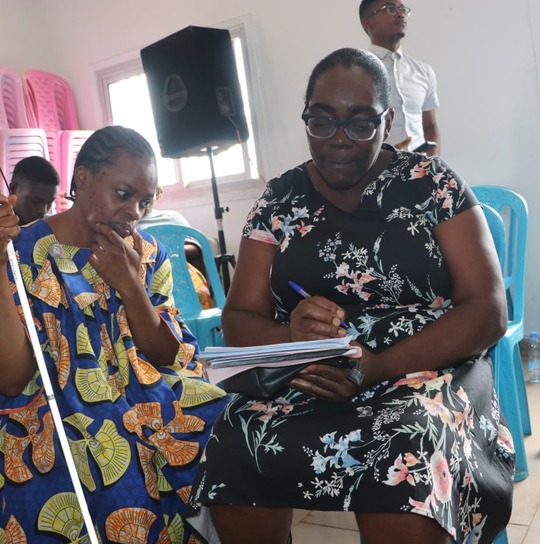 A blind woman holding a white cane sits beside a colleague writing notes on her behalf at a workshop.