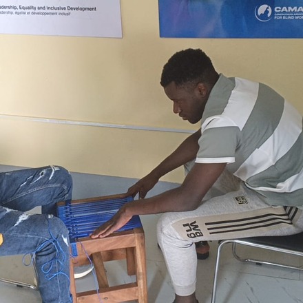 Two men weaving a stool seat with blue cord at the CAMAB Centre, CAMAB banners on the wall behind them.