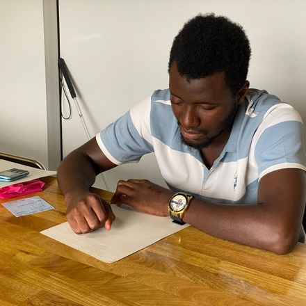 A learner writing Braille on a slate at a wooden table.
