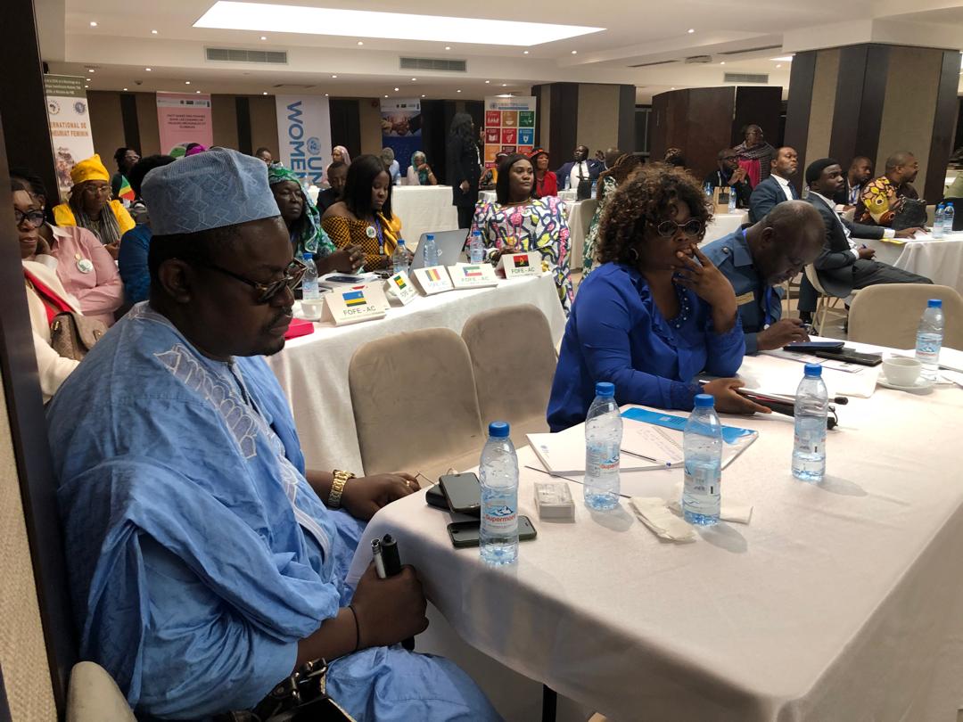 Conference room with 'WOMEN' banners; delegates seated at draped tables, a man in a blue boubou and cap in the foreground.