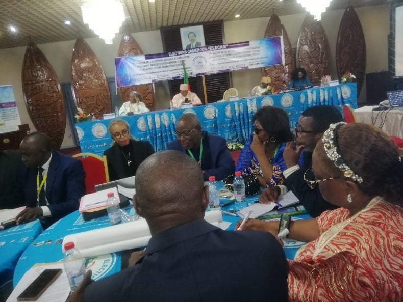 Formal conference session; delegates around a large blue-draped table, head table and national crest behind.