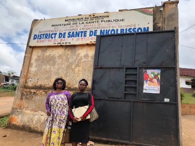 Two women stand at the gate of a compound; the wall sign reads 'Republique du Cameroun - Ministere de la Sante Publique - District de Sante de Nkolbisson'.