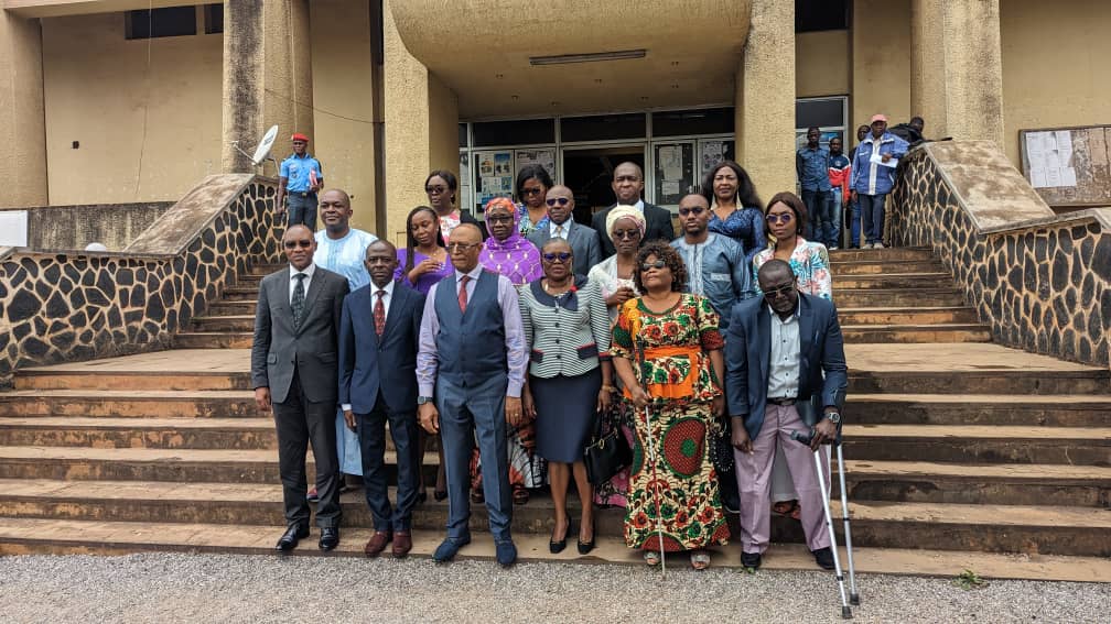 Formal group photograph on the steps of a public building: about fourteen people, officials in suits, one woman on crutches, a police officer at the left.