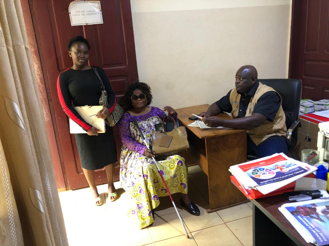 Office interview: a woman in dark glasses seated with a bag, a man at a desk taking notes, a second woman standing.