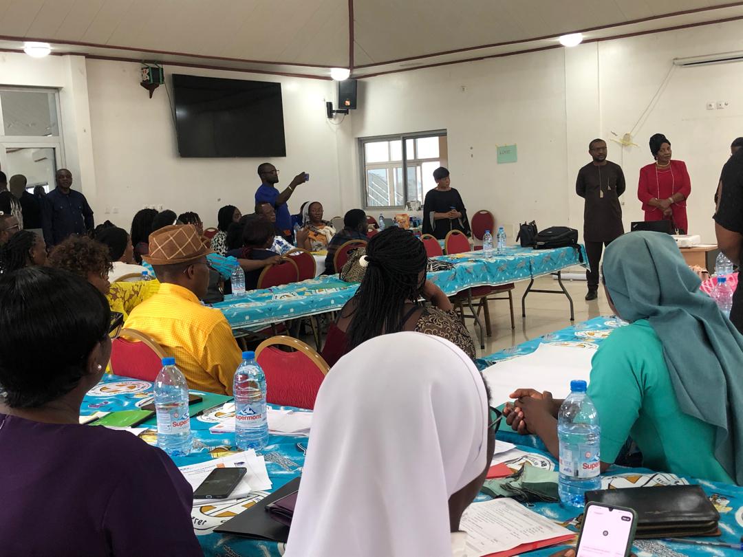 Conference room from the back; many participants at draped tables, speakers standing at the front.